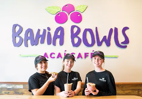 Three smiling team members at Bahia Bowls Acai Café stand in front of the restaurant’s logo, each holding colorful acai bowls and smoothies, representing the brand’s fresh, vibrant, and healthy food concept.